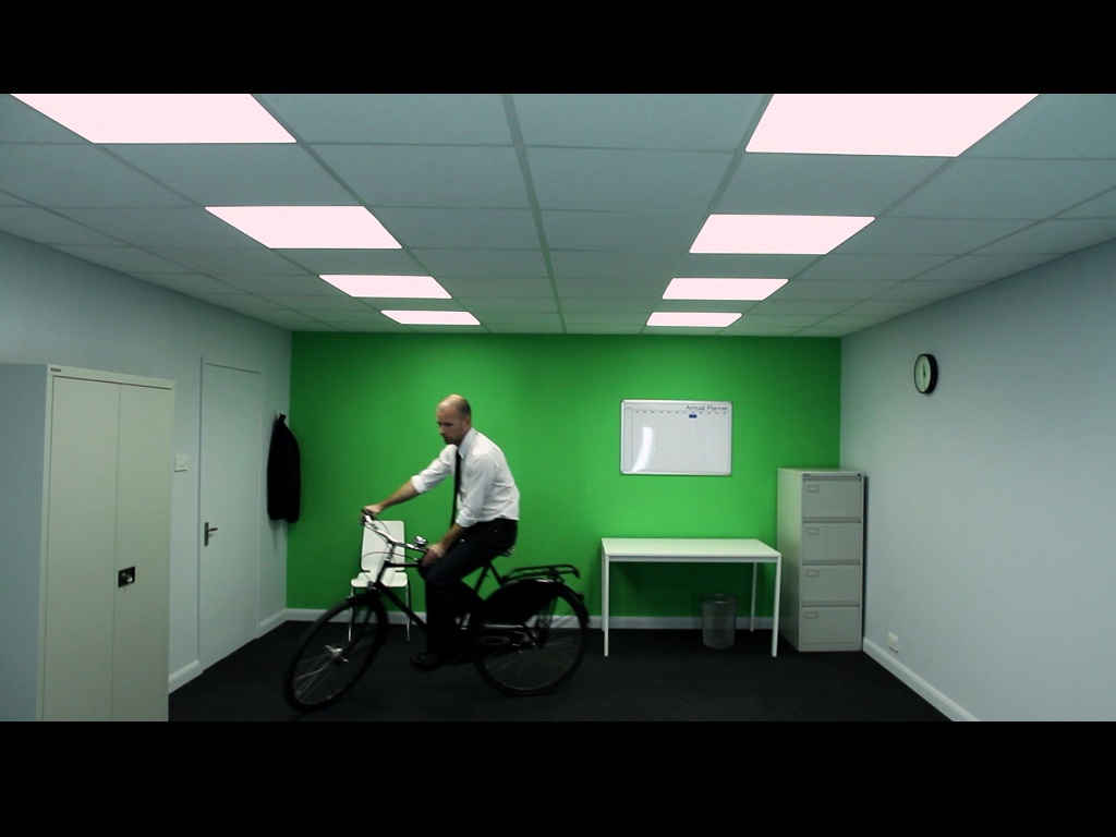 Kunst im Sommer 2025 in Göppingen: Ein Mann im weißen Hemd und schwarzer Krawatte fährt auf einem Fahrrad durch ein minimalistisch möbliertes Büro mit grün gestrichener Rückwand – Szene aus dem Video 10x10 von John Wood & Paul Harrison.
