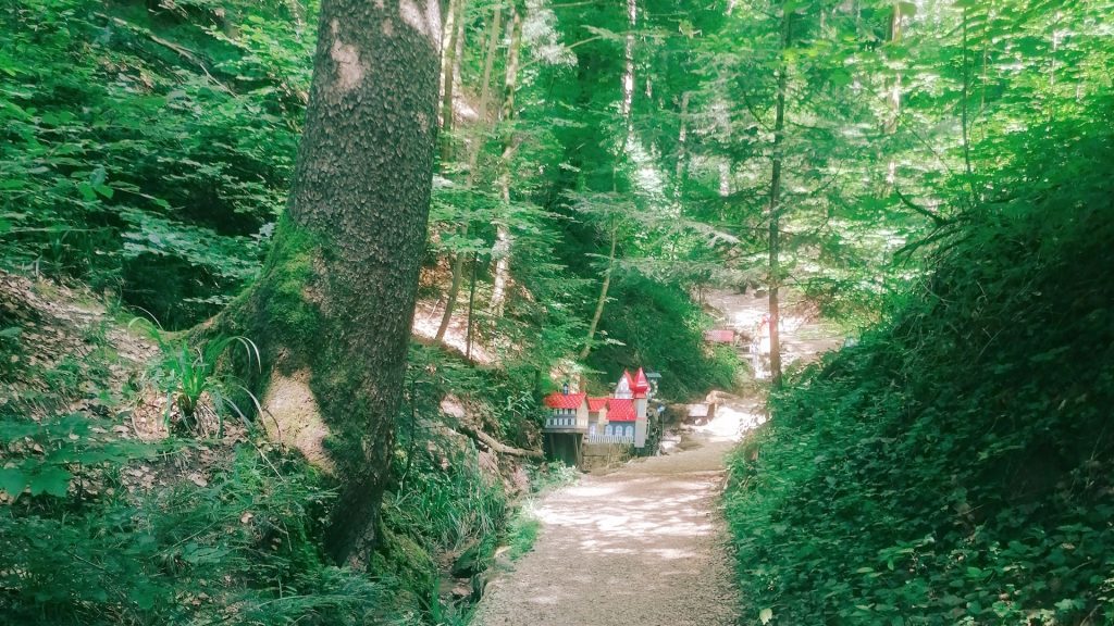 Ein idyllischer Waldweg führt durch dichtes Grün in Richtung Schelmenklinge in Lorch, wo zwischen den Bäumen kleine, wasserbetriebene Modelle mit roten Dächern sichtbar werden.