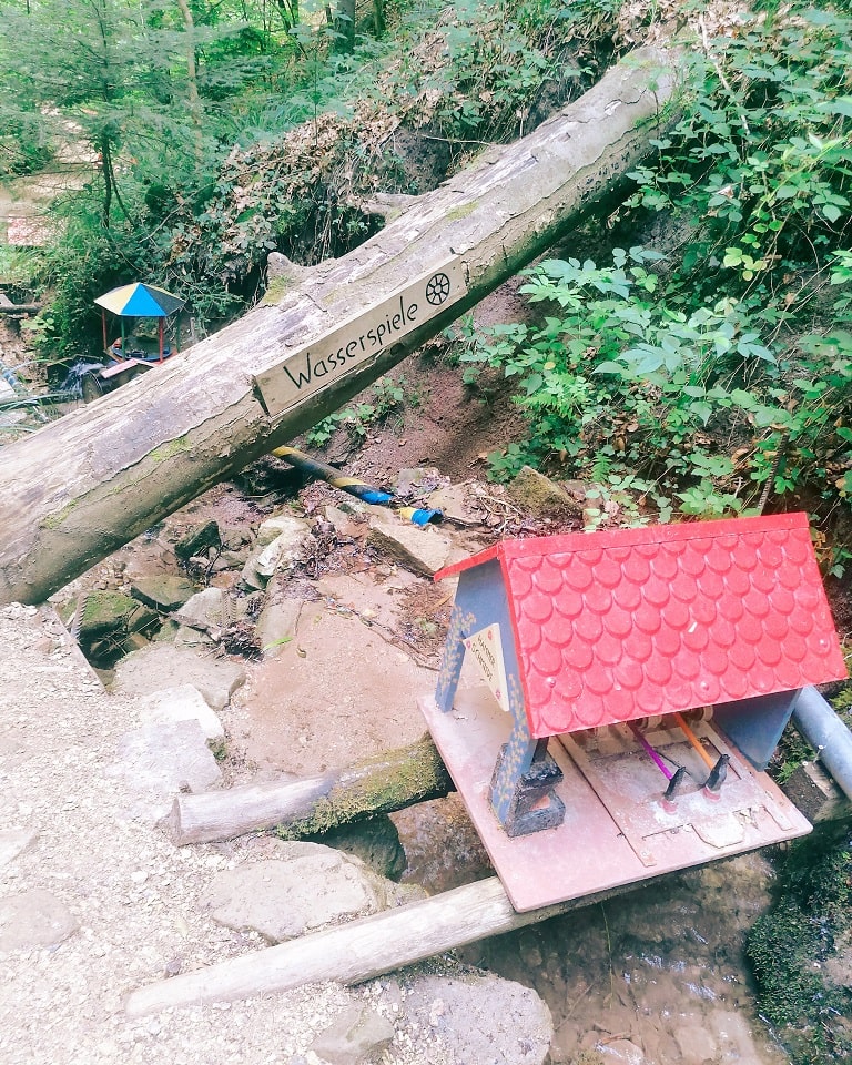 Ein umgestürzter Baum mit einem Holzschild „Wasserspiele“ markiert eine weitere Station der Schelmenklinge in Lorch, wo ein kleines Hausmodell über einem Bachlauf montiert ist.