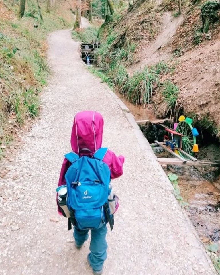 Ein Kind mit pinker Jacke und blauem Rucksack läuft einen Waldweg entlang, neben dem ein buntes Wasserrad vom Bach angetrieben wird – eine der vielen Mitmachstationen der Schelmenklinge in Lorch.