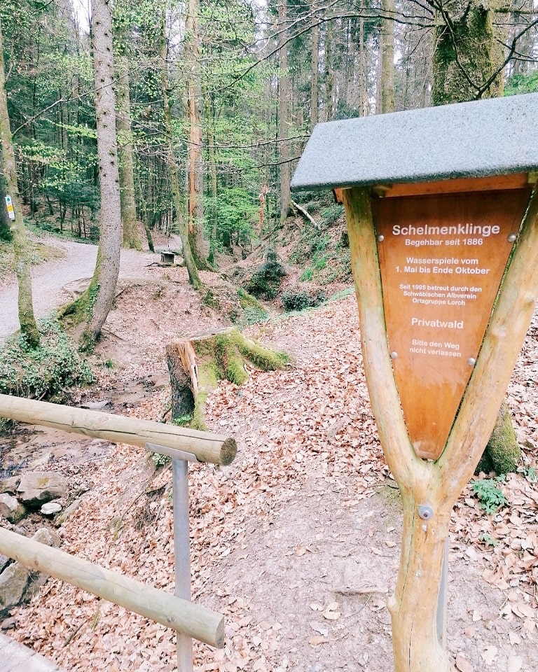 Holzschild am Waldrand informiert über die Schelmenklinge in Lorch – begehbar seit 1886 und mit Wasserspielen von Mai bis Oktober – umgeben von Bäumen und Laub.