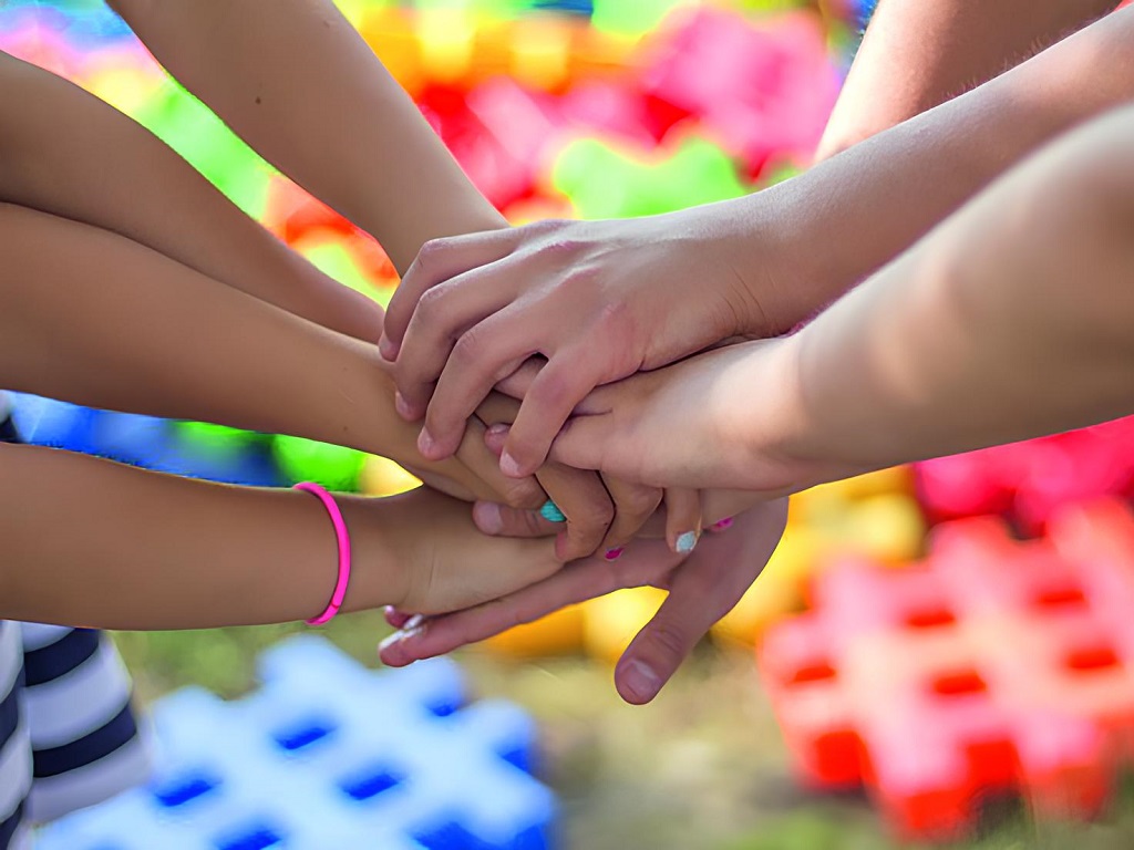 Mehrere Kinder legen ihre Hände in der Mitte übereinander – ein Zeichen von Teamgeist und Gemeinschaft, aufgenommen vor einem bunten Hintergrund im Freien.