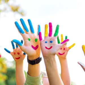 Kinderhände mit bunten Farben bemalt, in die Luft gestreckt, jede Hand zeigt ein fröhliches Gesicht – ein kreativer Sommermoment voller Spaß und Farbe.