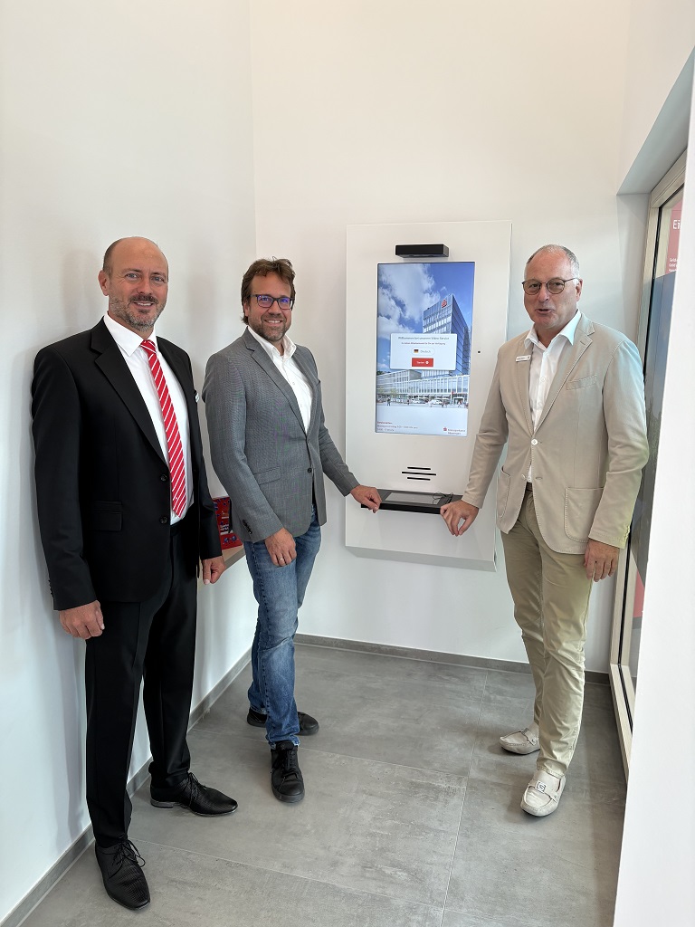 Drei Männer in Anzügen stehen lächelnd neben der neuen digitalen Videostele der Kreissparkasse Wäschenbeuren, die an einer weißen Wand montiert ist.