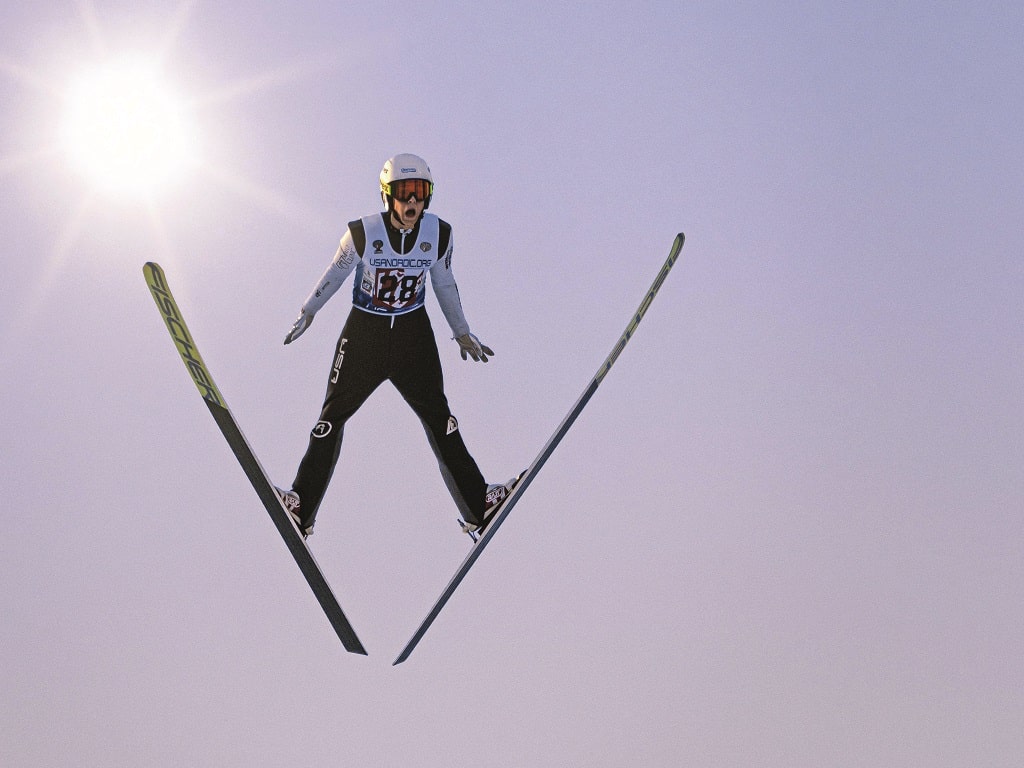 Eine Skispringerin in voller Ausrüstung schwebt in der Luft vor blauem Himmel und strahlender Sonne – Sommertraining auf der Mattenschanze beim Schülerferienprogramm Süßen 2025