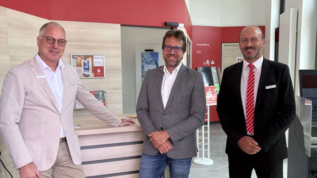 Drei Männer in Anzügen stehen in der modernen, rot-weißen Filiale in Wäschenbeuren der Kreissparkasse Göppingen und lächeln in die Kamera. Im Hintergrund ist die neue Videostele zu sehen.