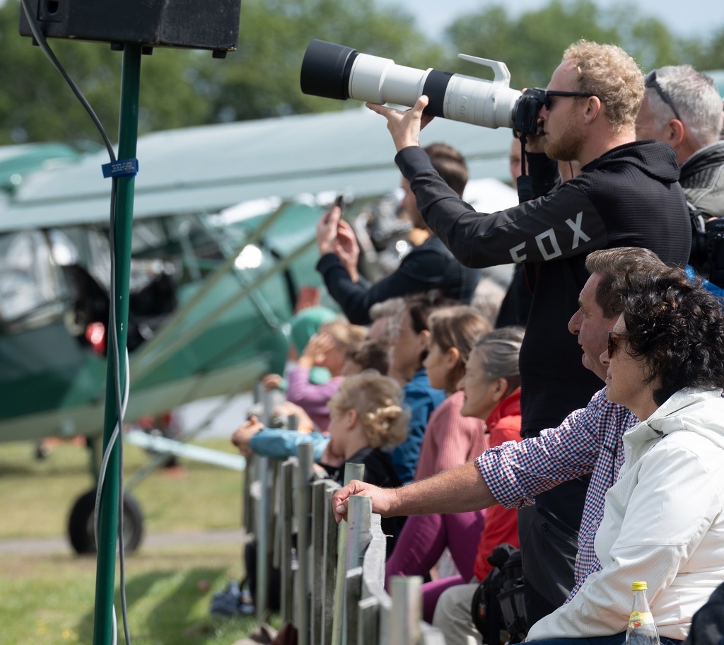 Familien stehen und sitzen am Zaun eines Flugplatzes, schauen gespannt in den Himmel; im Vordergrund ein Fotograf mit Teleobjektiv, im Hintergrund ein grünes Oldtimerflugzeug.