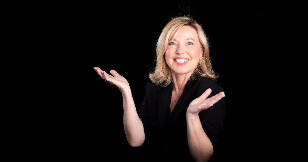 Kabarettistin Simone Solga mit ausgebreiteten Armen und strahlendem Lächeln vor schwarzem Hintergrund