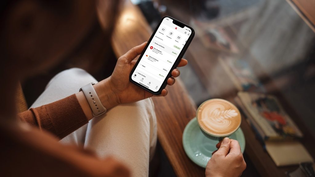 Hand hält Smartphone mit Sparkassen-App und integriertem Finanzplaner, daneben Cappuccino auf einem Tisch.