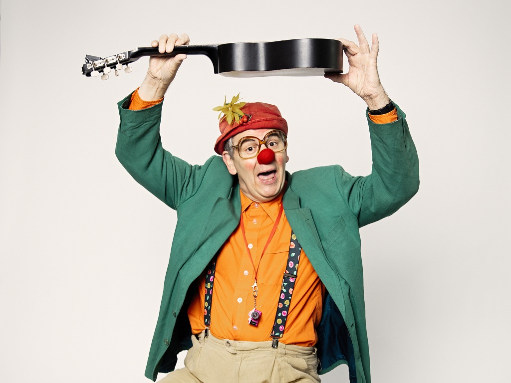 Clown Ludger Hoffkamp mit roter Nase, grüner Jacke und Gitarre, bekannt für seinen Vortrag "Humor als Türöffner für Menschen mit Demenz".