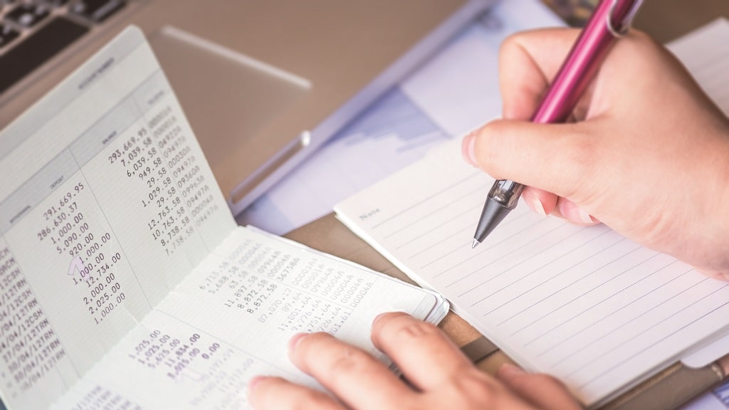 Hand schreibt Zahlen aus einem Kontoauszug in ein Notizbuch. Früher Haushaltsbuch, heute digital mit dem Finanzplaner.