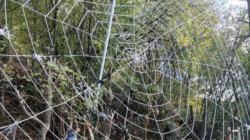 Großes Spinnennetz mit schwarzen und weißen Spinnen als Halloween-Dekoration im Freien, vor herbstlich bunten Bäumen.