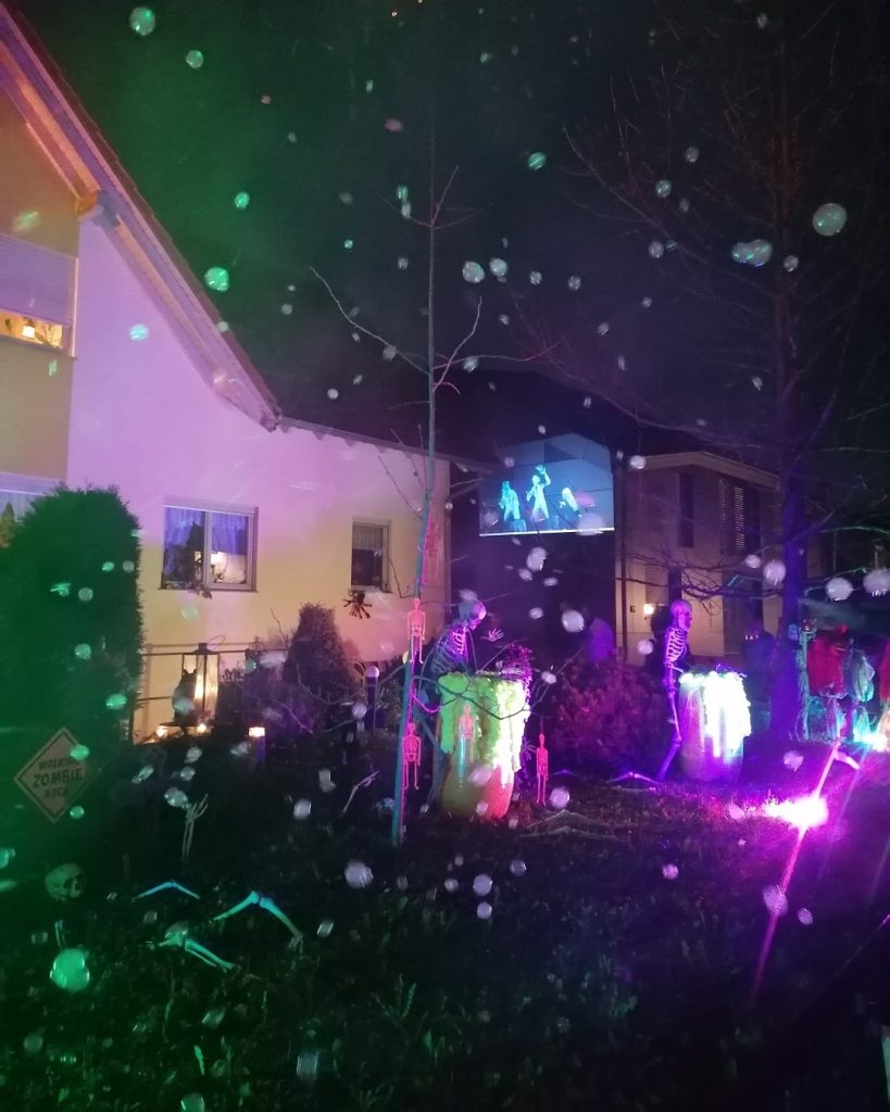 Leuchtend geschmückter Gruselgarten in Bad Boll bei Nacht mit Nebel, bunten Lichtern, Skeletten und Halloween-Projektion auf Hauswand.