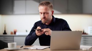 Eine Person sitzt am Schreibtisch vor einem Laptop und fotografiert ein Dokument mit dem Smartphone, um eine Rechnung digital zu erfassen.