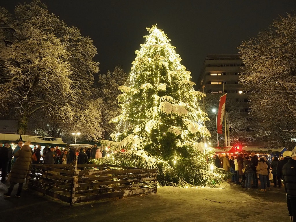 Groß beleuchteter Weihnachtsbaum auf dem Holzheimer Weihnachtsmarkt, umgeben von Besuchern und verschneiten Bäumen bei Nacht.