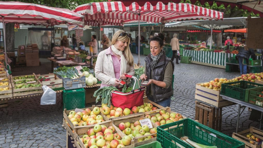 Zwei Personen bezahlen auf einem Wochenmarkt kontaktlos mit der Sparkassen-Card; frisches Obst und Gemüse in Holzkisten im Vordergrund.