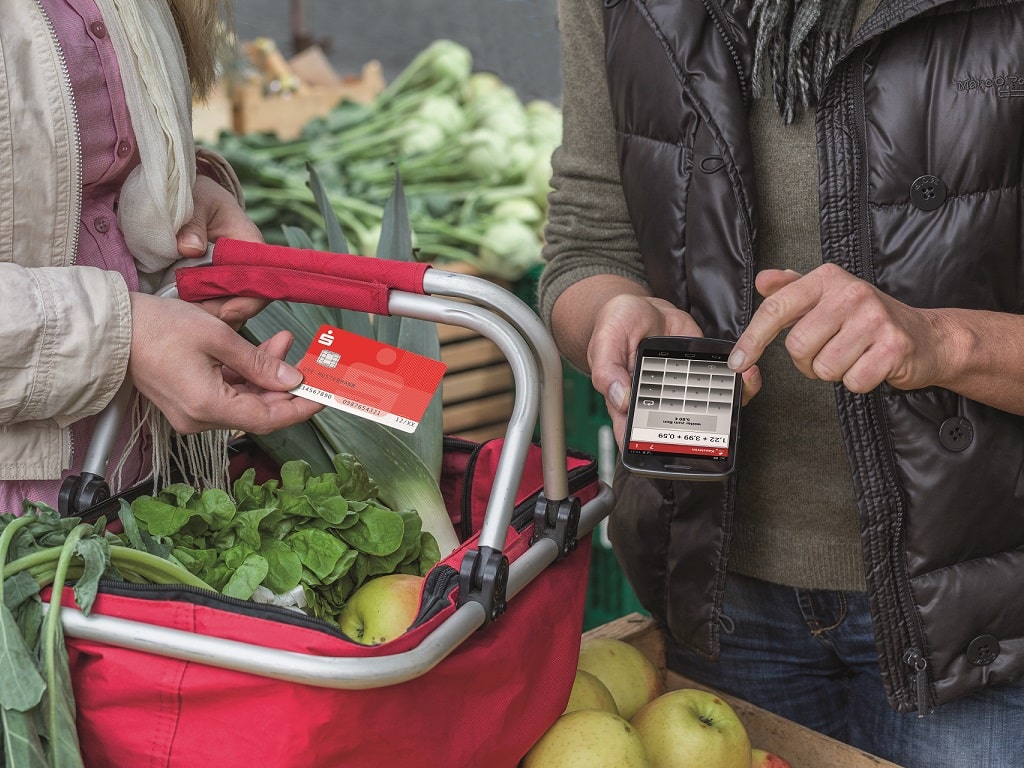 Zwei Personen auf dem Wochenmarkt zeigen eine Sparkassen-Card und eine Bezahl-App auf dem Smartphone; ein Einkaufskorb mit frischem Gemüse im Vordergrund