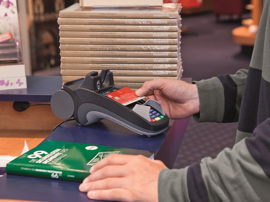 Person bezahlt kontaktlos mit der Sparkassen-Card an einem Kartenterminal in einem Geschäft; ein Buch liegt auf dem Tresen.
