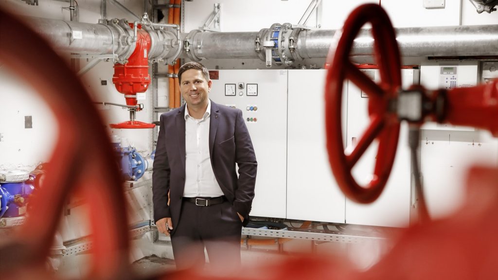 Alexander Lange, Geschäftsführer von FGM Gebäudemanagement, steht in einem Technikraum mit Rohrleitungen und großen roten Ventilrädern.