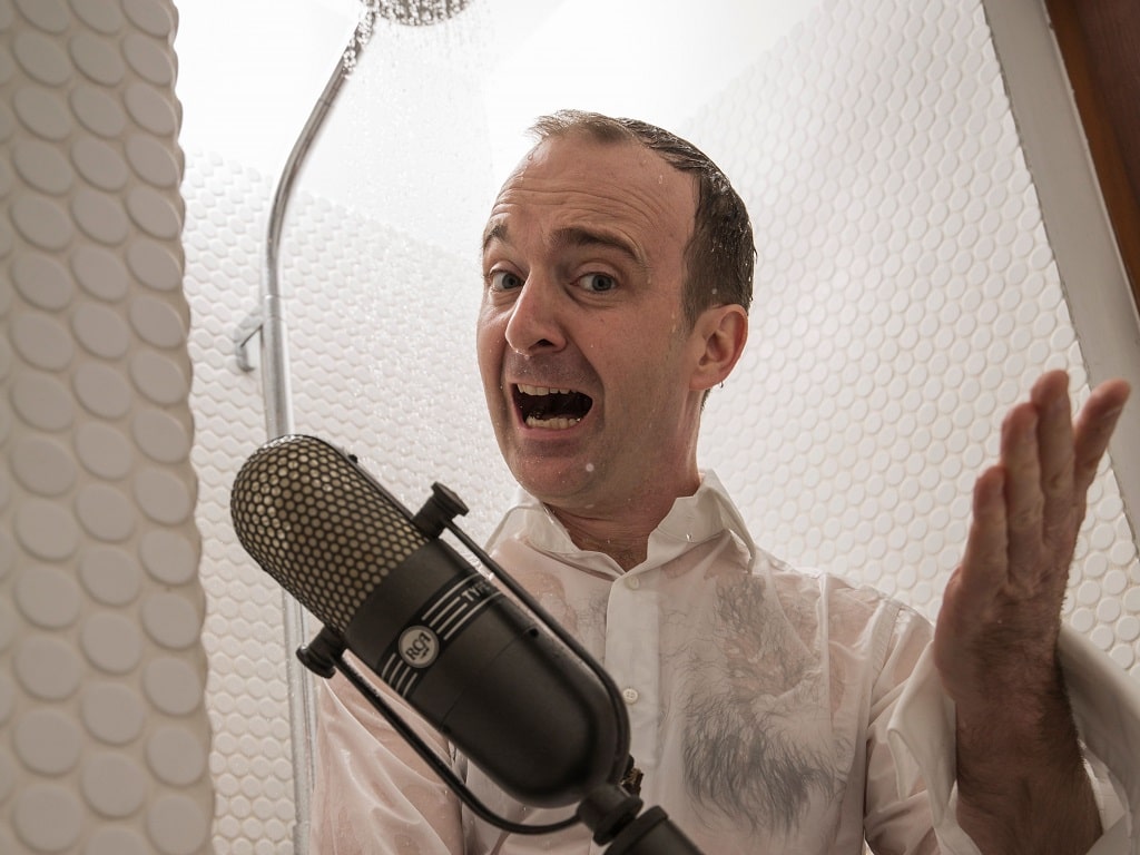 Musiker Patrick Bopp singt ins Mikrofon und steht humorvoll unter der Dusche – Symbol für das Mitsingkonzert „Aus voller Kehle für die Seele“ in Göppingen.