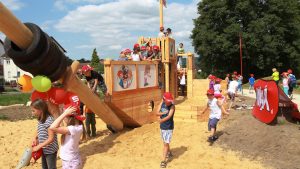 Kinder spielen auf einem großen KNAX-Spielschiff aus Holz auf einem Sandspielplatz. Viele Kinder tragen rote Kappen und bewegen sich fröhlich rund um das Kletterschiff. KNAX-Illustrationen sind an der Holzwand angebracht, daneben stehen bunte Luftballons und ein KNAX-Banner. Im Hintergrund befinden sich Bäume und Wohnhäuser.