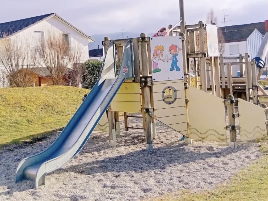 Holz-Kletterschiff mit blauer Rutsche auf dem KNAX-Spielplatz in Heiningen. Die Konstruktion ist mit KNAX-Illustrationen verziert und steht auf Kies. Im Hintergrund befinden sich Wohnhäuser und eine leicht hügelige Grünfläche.