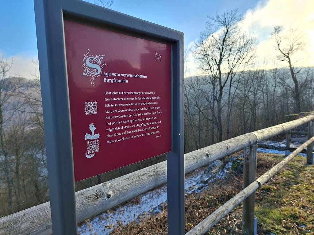 Informationstafel mit der Sage vom verwunschenen Burgfräulein an der Burgruine Hiltenburg, aufgestellt am Aussichtspunkt mit Blick über den winterlichen Wald.