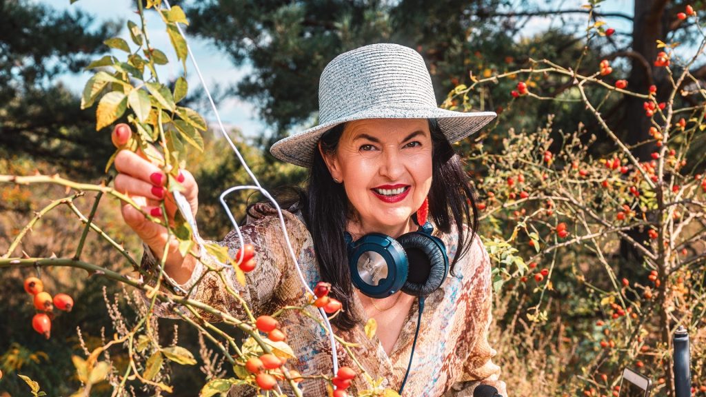 Titelbild für den Beitrag "Pflanzenmusik in der Ausstellung Furry Flowers". Person im Freien mit Kopfhörern zwischen Pflanzen, hält einen Zweig und arbeitet mit Pflanzenmusik-Equipment.