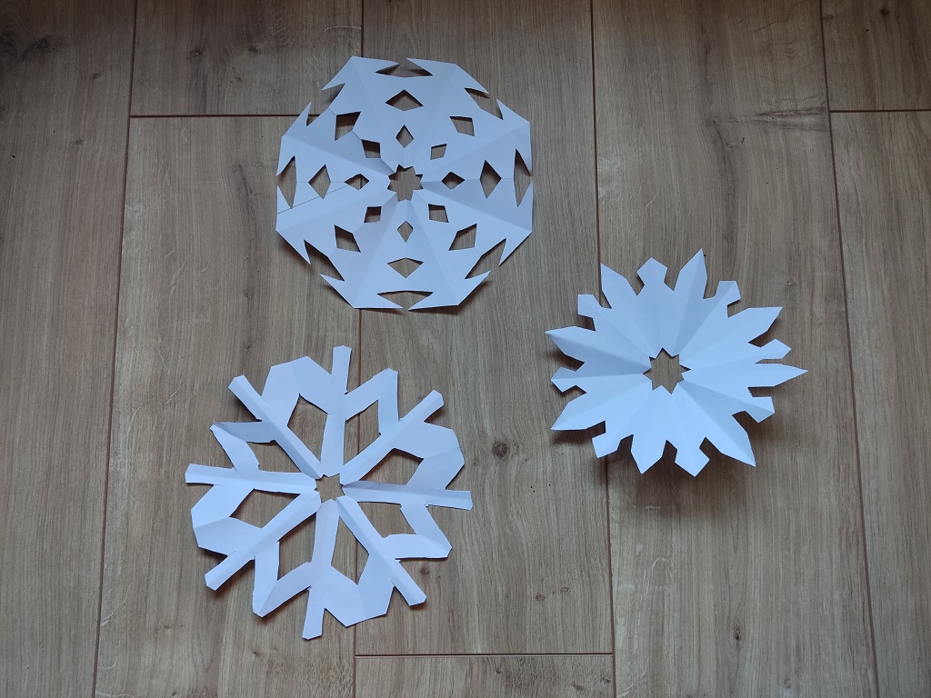 Drei unterschiedlich geschnittene Schneeflocken aus weißem Papier liegen auf einem Holzboden.