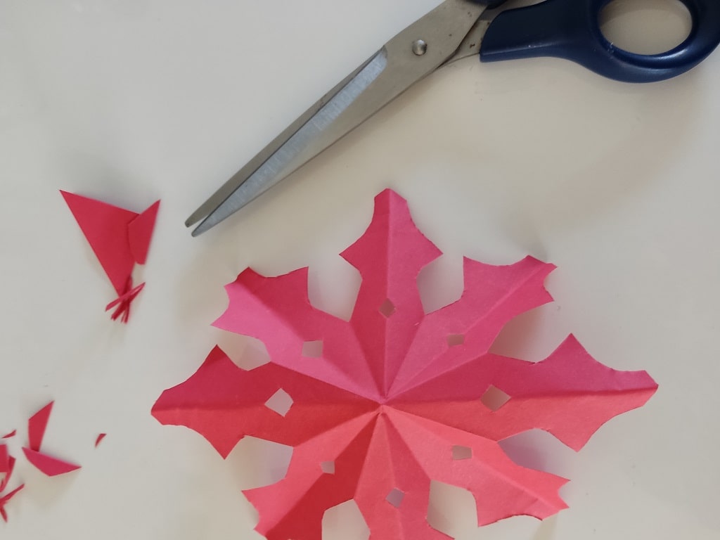 Aufgefaltete Papier-Schneeflocke aus dem Schneeflocken-Scherenschnitt liegt auf dem Tisch, daneben eine Schere und Papierreste.