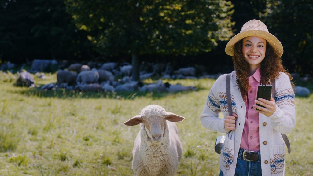 Frau mit Smartphone steht auf einer Wiese neben einem Schaf und symbolisiert digitalen Finanzüberblick mit Multibanking