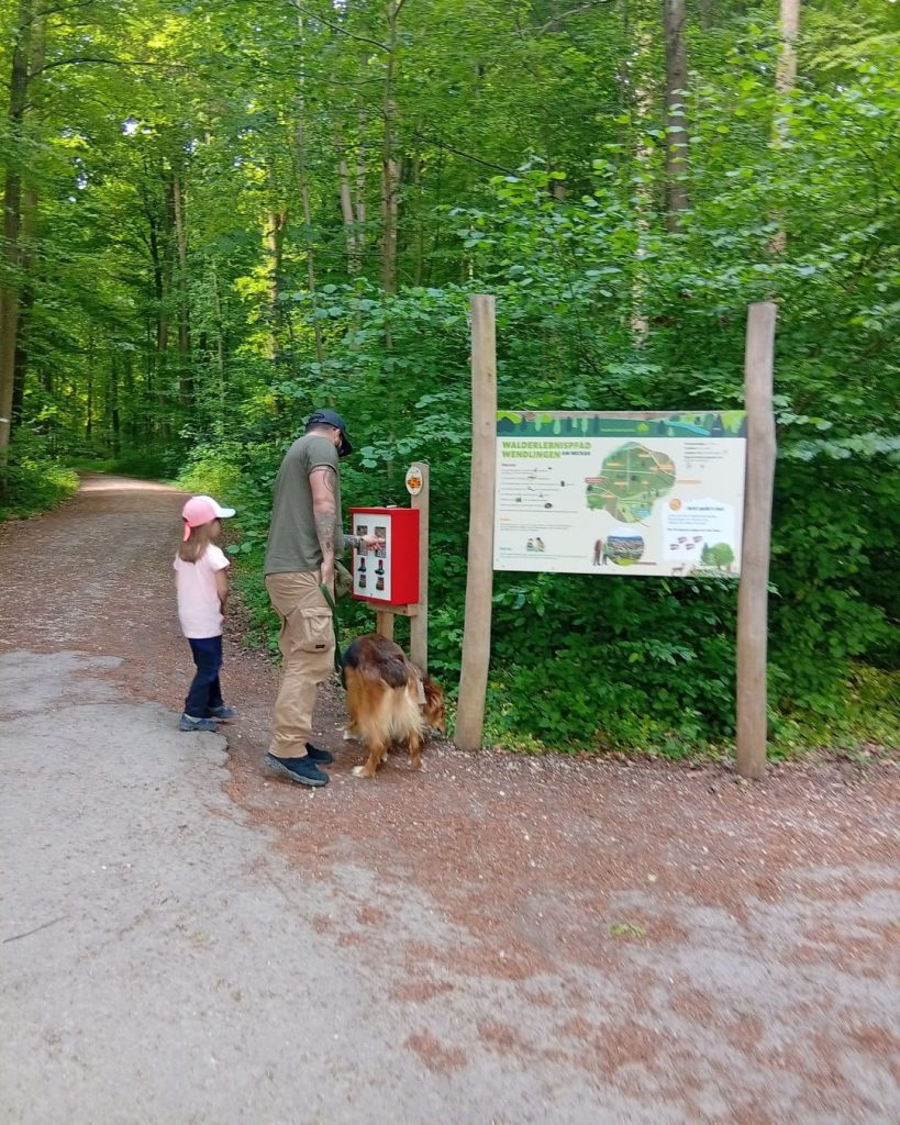 Vater und Kind kaufen Holzkugeln am Automaten. Daneben eine Informationstafel im grünen Wald.