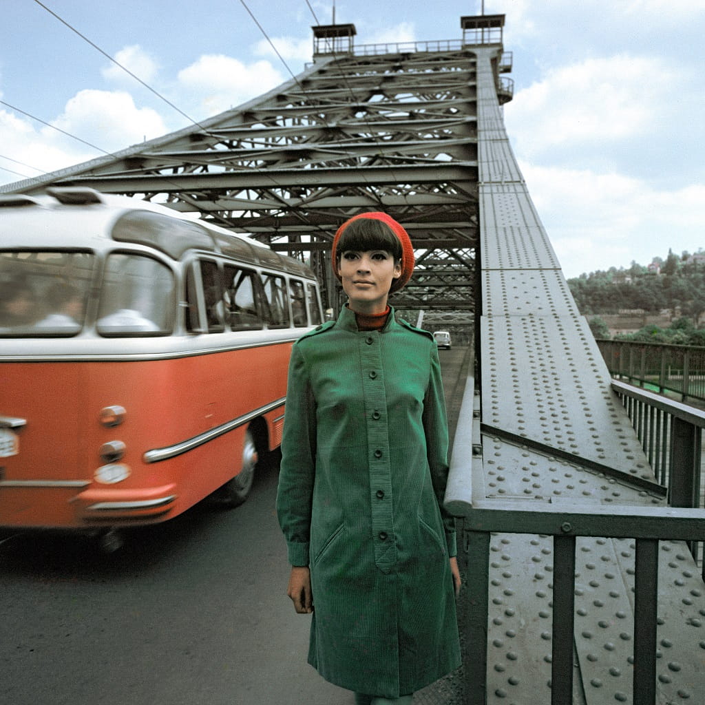 Farbige Fotografie von Arno Fischer: Frau in grünem Mantel steht auf einer Brücke, im Hintergrund fährt ein Bus vorbei, Aufnahme aus dem Jahr 1967.