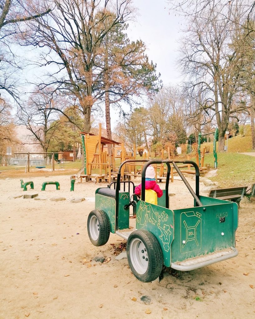 Kind spielt auf einem grünen Spielauto. Im Hintergrund Holzspielgeräte und hohe Bäume.