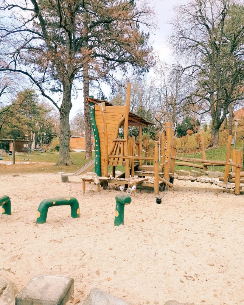 Holzspielanlage mit Rutsche und Balancier-Elementen im Sandbereich des Spielplatzes im Stadtpark Geislingen, umgeben von großen Bäumen.