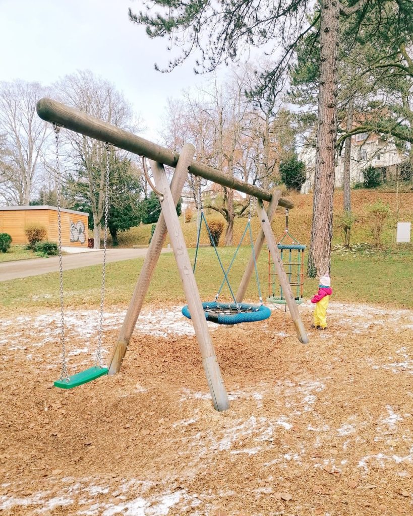 Schaukelanlage mit Brettschaukel und Nestschaukel. Ein Kind steht daneben auf der Rasenfläche.