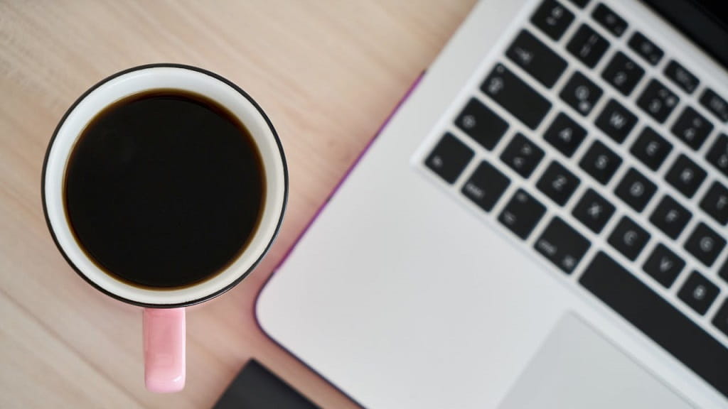 Kaffeetasse neben einem Laptop auf einem Tisch, aufgenommen von oben. Symbolbild für die Suche nach Kurzanleitungen im Internet.