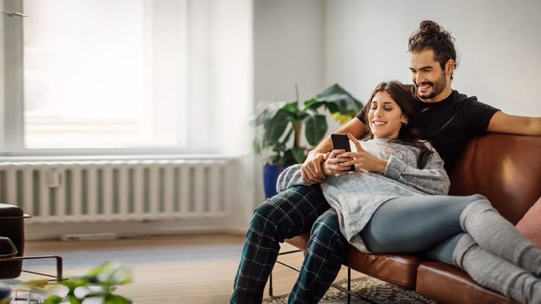 Paar sitzt entspannt auf dem Sofa und schaut auf ein Smartphone, Symbolbild für Kurzanleitungen und Hilfe beim Online-Banking.