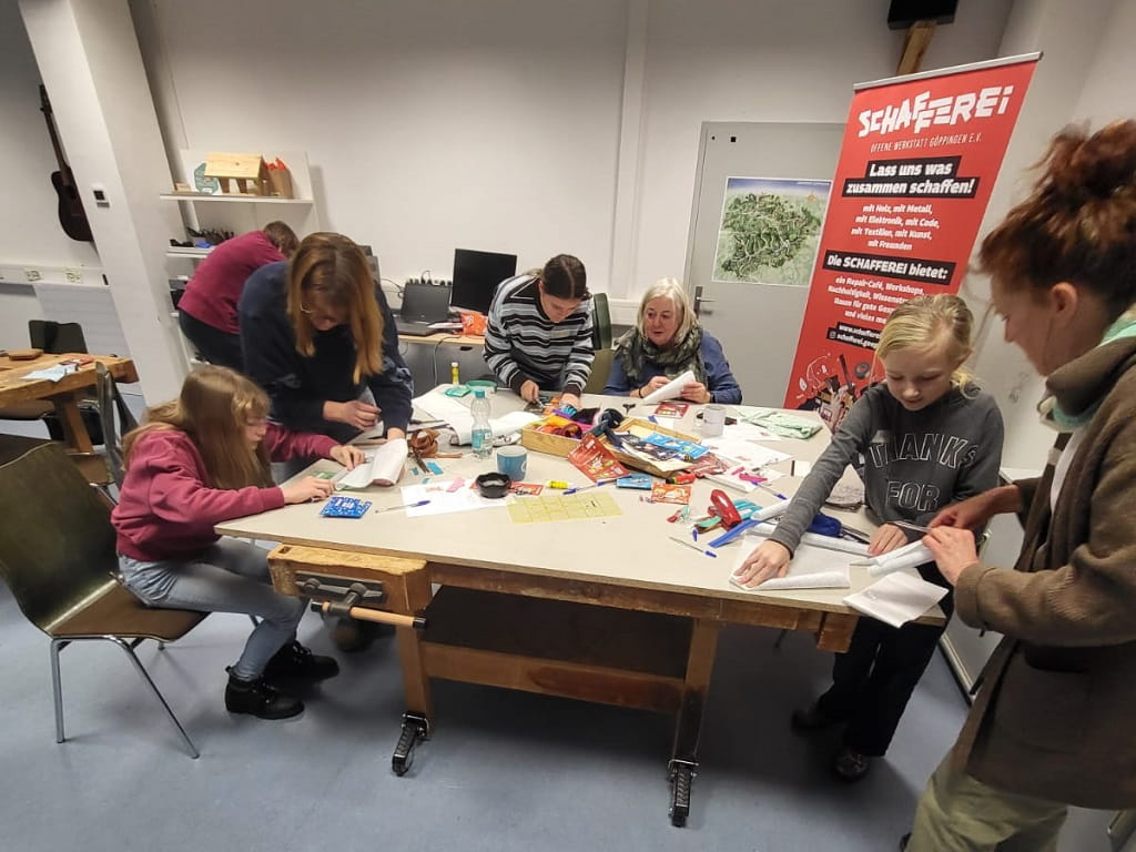 Teilnehmende eines Upcycling-Workshops arbeiten gemeinsam an einem Tisch und gestalten aus Schokoladenverpackungen neue Produkte.