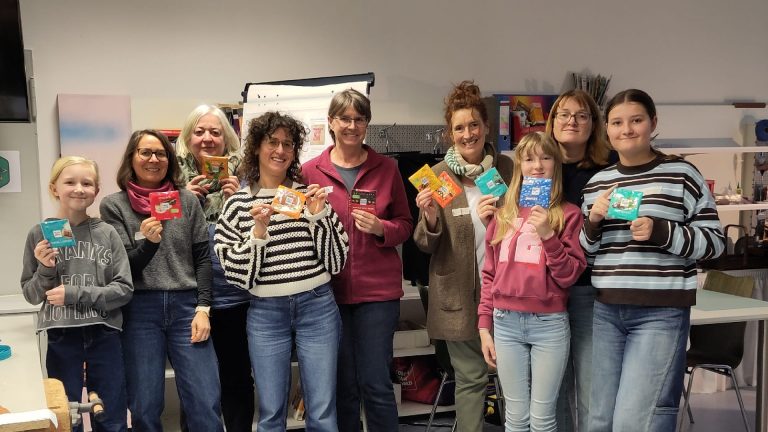 Teilnehmende eines Upcycling-Workshops in der SCHAFFEREI Göppingen stehen gemeinsam in einer Werkstatt und halten selbst gestaltete Geldbeutel aus Ritter-Sport-Verpackungen in die Kamera.