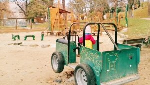 Kleinkind spielt auf einem grünen Spielauto im Sandbereich des Spielplatzes im Stadtpark Geislingen, im Hintergrund Holzspielgeräte und Bäume.