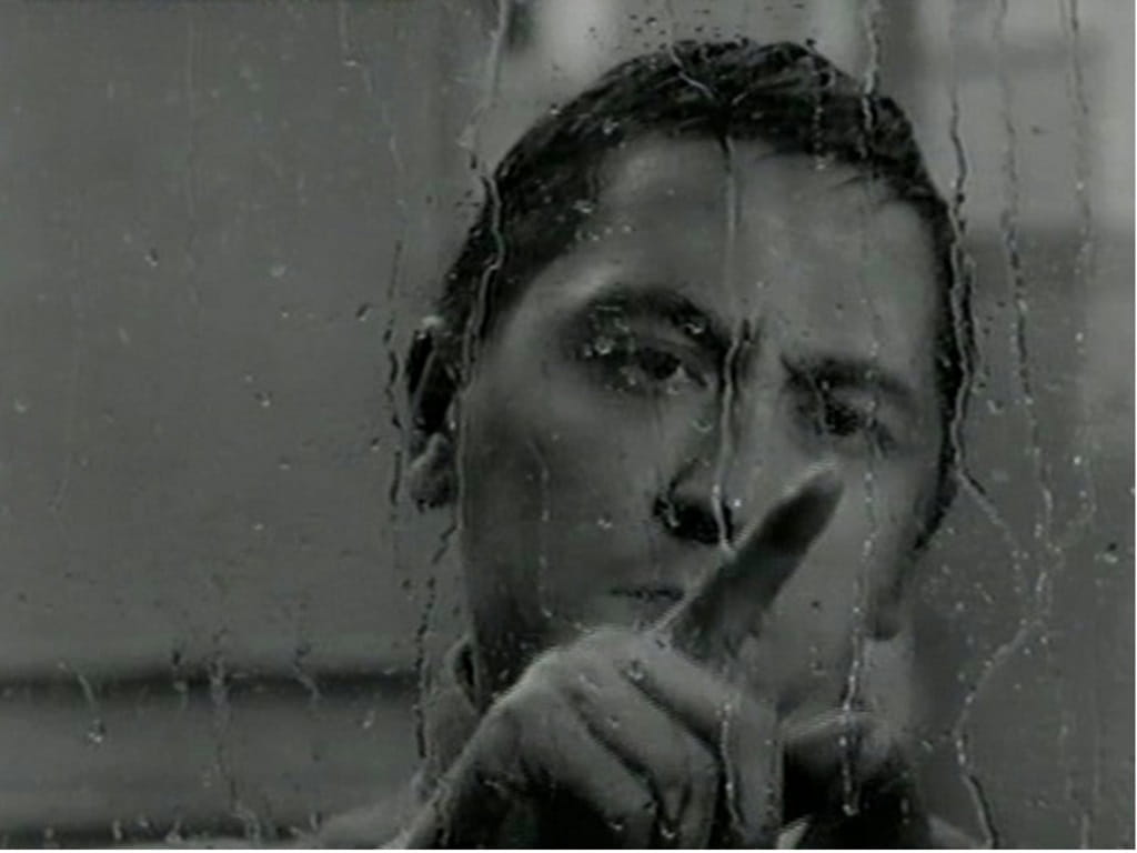 Schwarzweiß-Filmstill aus „Jonas“: Nahaufnahme eines Mannes hinter einer regennassen Scheibe, der mit dem Finger nach vorne zeigt.