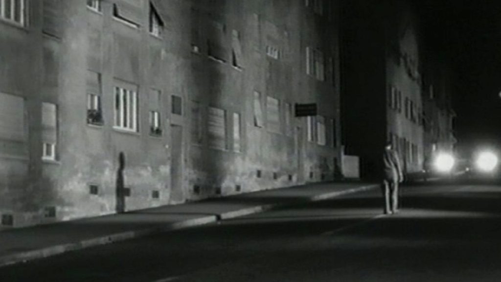 Schwarzweiß-Filmstill aus „Jonas“: Ein Mann steht nachts allein auf einer Straße zwischen hohen Wohnhäusern, Scheinwerferlicht im Hintergrund.