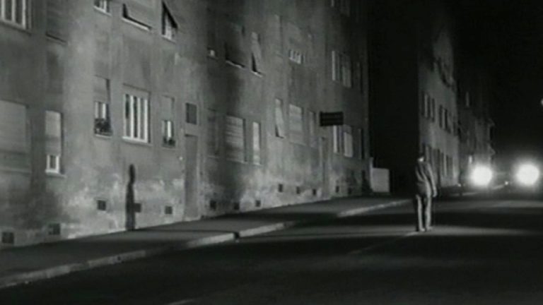 Schwarzweiß-Filmstill aus „Jonas“: Ein Mann steht nachts allein auf einer Straße zwischen hohen Wohnhäusern, Scheinwerferlicht im Hintergrund.