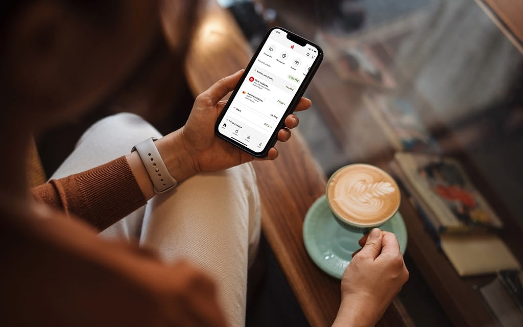 Person nutzt Smartphone mit Banking-App und erledigt mobiles Bezahlen bei einer Tasse Kaffee und gewinnt dabei mehr digitales Vertrauen