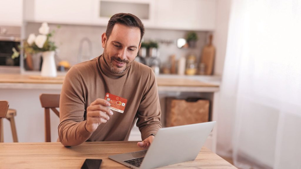 Mann sitzt am Tisch mit Laptop und hält eine Bankkarte in der Hand, während er Online-Banking nutzt und sich über digitales Vertrauen informiert