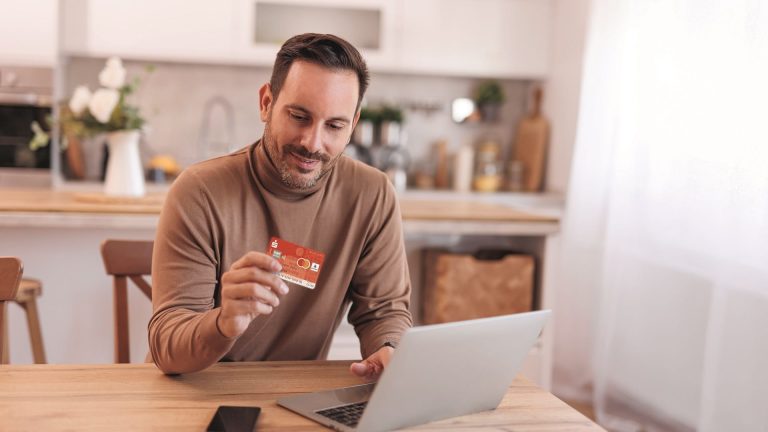Mann sitzt am Tisch mit Laptop und hält eine Bankkarte in der Hand, während er Online-Banking nutzt und sich über digitales Vertrauen informiert