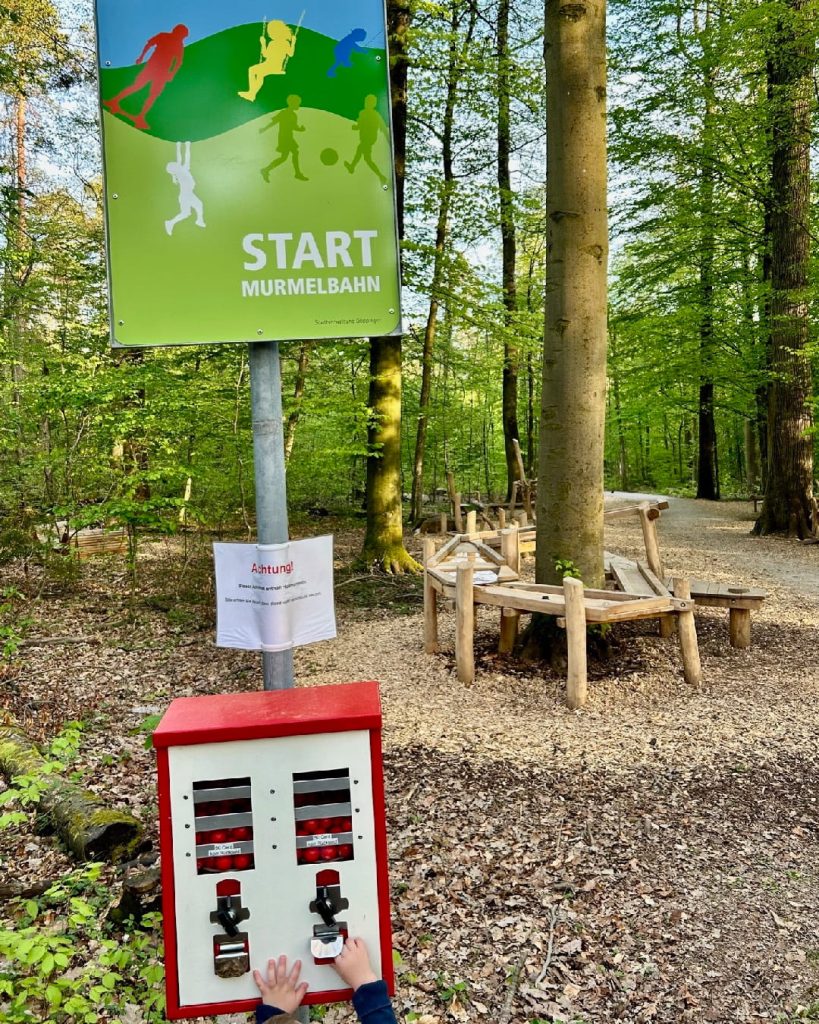 Startschild einer Murmelbahn im Wald mit Kugelautomat und Holzbahn im Hintergrund zum Thema Murmel wird olympisch