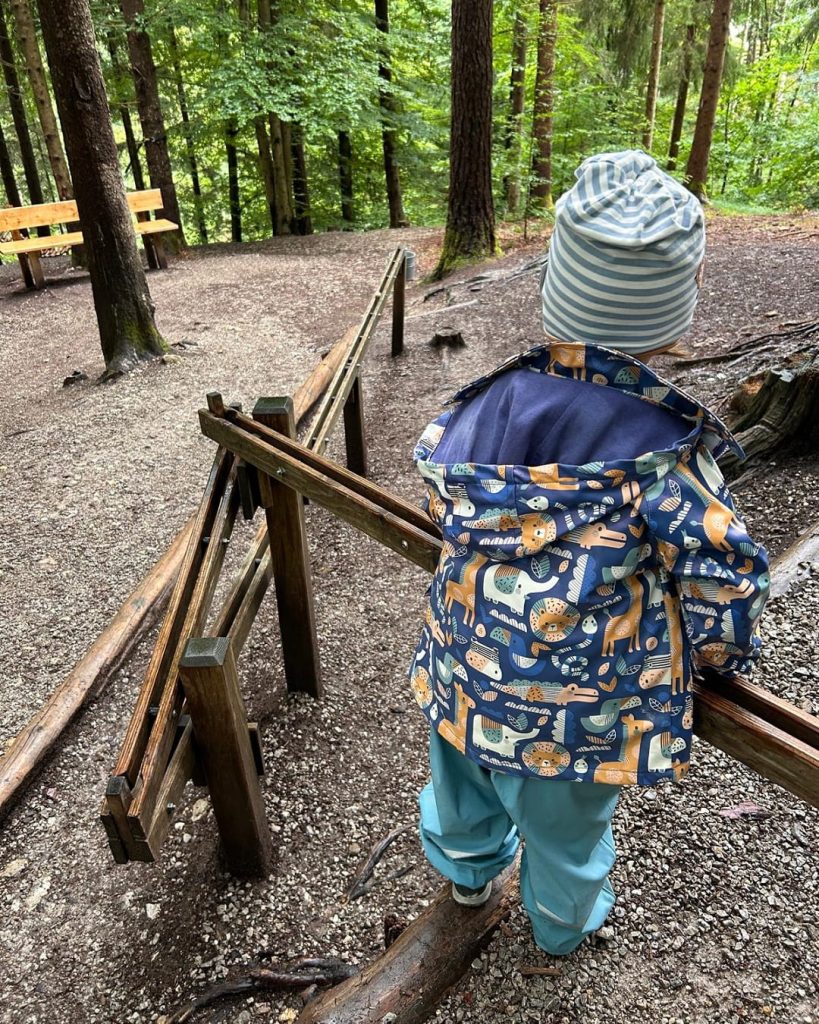 Kind mit Mütze und Jacke steht an einer Holz-Kugelbahn im Wald, im Hintergrund Bäume und Sitzbank