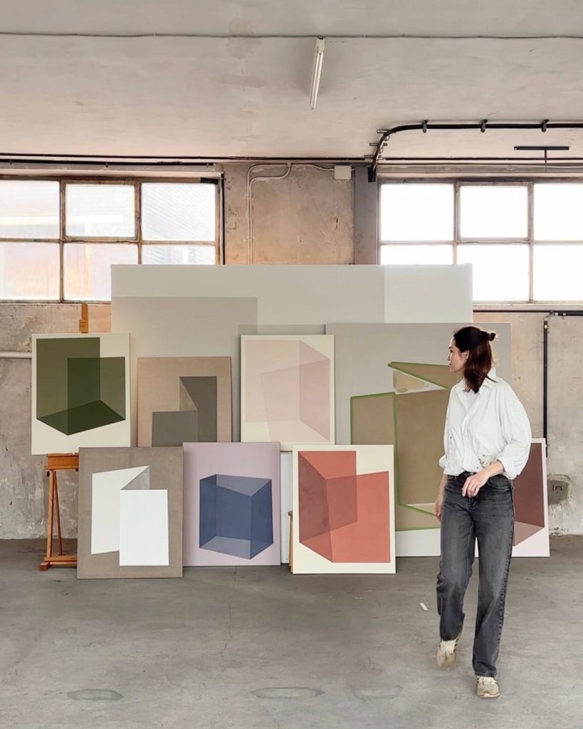 Carolin Kreutzer steht in ihrem Atelier vor mehreren großformatigen, geometrisch-abstrakten Gemälden.